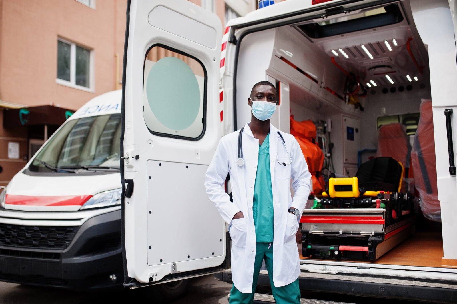 African male paramedic in face protective medical mask standing in front of ambulance car.